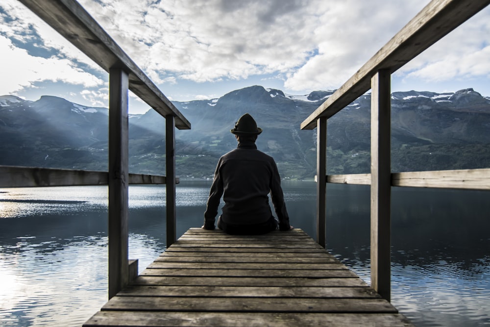 man in black hoodie sitting on wooden dock during daytime