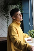 A person wearing a yellow sweatshirt is standing by a window with a cheerful expression. The background includes white tiles and green plants, providing a peaceful, natural setting.