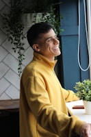 A person wearing a yellow sweatshirt is standing by a window with a cheerful expression. The background includes white tiles and green plants, providing a peaceful, natural setting.