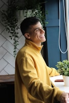 A person wearing a yellow sweatshirt is standing by a window with a cheerful expression. The background includes white tiles and green plants, providing a peaceful, natural setting.