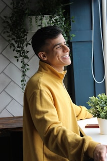 A person wearing a yellow sweatshirt is standing by a window with a cheerful expression. The background includes white tiles and green plants, providing a peaceful, natural setting.