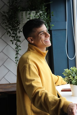 A person wearing a yellow sweatshirt is standing by a window with a cheerful expression. The background includes white tiles and green plants, providing a peaceful, natural setting.