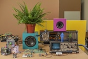 A table cluttered with a variety of electronic components and decorative items. A potted fern sits atop a blue speaker, next to a smaller purple speaker. A vintage oscilloscope is surrounded by wires and circuit boards. A small blue figurine stands next to a black potted plant. In the foreground, colorful electronic projects are strewn across the surface.