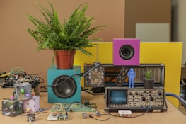 A table cluttered with a variety of electronic components and decorative items. A potted fern sits atop a blue speaker, next to a smaller purple speaker. A vintage oscilloscope is surrounded by wires and circuit boards. A small blue figurine stands next to a black potted plant. In the foreground, colorful electronic projects are strewn across the surface.