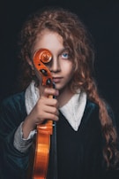 A young person with curly auburn hair holds a violin upright, covering one eye with the scroll. They wear a dark velvet garment with a white lace collar. The background is dark, emphasizing their bright blue eyes and serious expression.