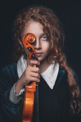 A young person with curly auburn hair holds a violin upright, covering one eye with the scroll. They wear a dark velvet garment with a white lace collar. The background is dark, emphasizing their bright blue eyes and serious expression.