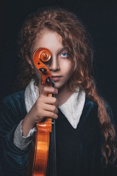 A young person with curly auburn hair holds a violin upright, covering one eye with the scroll. They wear a dark velvet garment with a white lace collar. The background is dark, emphasizing their bright blue eyes and serious expression.