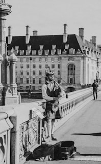 A man in traditional Scottish attire is playing the bagpipes on a bridge. The background features a large, historic building with numerous windows and chimneys. The bridge is adorned with ornate designs, and there is an open instrument case on the ground in front of him.