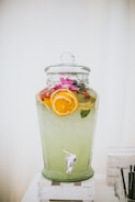 A large glass beverage dispenser filled with a light green drink, garnished with slices of oranges and lemons. The dispenser is topped with a glass lid and is placed on a white wooden stand. A tap is visible at the bottom for serving.
