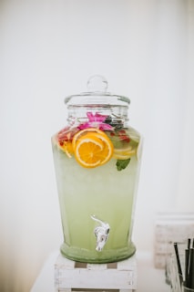 A large glass beverage dispenser filled with a light green drink, garnished with slices of oranges and lemons. The dispenser is topped with a glass lid and is placed on a white wooden stand. A tap is visible at the bottom for serving.