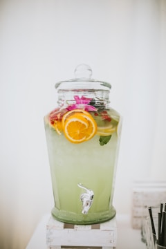 A large glass beverage dispenser filled with a light green drink, garnished with slices of oranges and lemons. The dispenser is topped with a glass lid and is placed on a white wooden stand. A tap is visible at the bottom for serving.