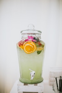 A large glass beverage dispenser filled with a light green drink, garnished with slices of oranges and lemons. The dispenser is topped with a glass lid and is placed on a white wooden stand. A tap is visible at the bottom for serving.