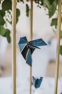 An intricately folded origami bird is suspended by a string between vertical wooden bars, against a blurred background of leaves. The origami is a deep blue with white edges, showcasing its artistic and delicate construction.