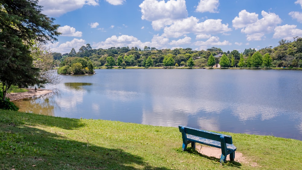青と白の曇り空の下、湖の近くの緑の芝生のフィールドに青い木製のベンチ