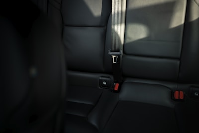 A close-up view of a car's back seat featuring black leather upholstery, red seatbelt latch buttons, and a fabric seatbelt. The lighting creates shadows and highlights on the surface.