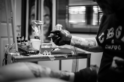 A person with tattooed arms is holding a tattoo machine while seated at a workstation. The workstation is equipped with various inks, a cup, and other tattooing supplies. The background features art pieces and window blinds, suggesting a tattoo studio environment.