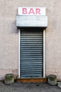 A closed rolling shutter door with a sign above it displaying the word 'BAR' in pink lettering. The structure is framed on a plain, muted wall, with two short concrete bollards on either side of the entrance.