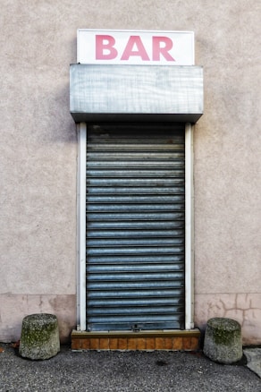 A closed rolling shutter door with a sign above it displaying the word 'BAR' in pink lettering. The structure is framed on a plain, muted wall, with two short concrete bollards on either side of the entrance.