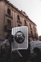 A protest scene with a hand holding a sign that repeats the phrase 'NO ES MI PRESIDENTE' alongside a stylized image of a man's face. The background features an old, grand building with arched windows and a crowd of people holding various signs, suggesting a demonstration in an urban environment.