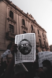 A protest scene with a hand holding a sign that repeats the phrase 'NO ES MI PRESIDENTE' alongside a stylized image of a man's face. The background features an old, grand building with arched windows and a crowd of people holding various signs, suggesting a demonstration in an urban environment.