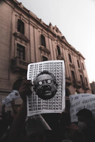 A protest scene with a hand holding a sign that repeats the phrase 'NO ES MI PRESIDENTE' alongside a stylized image of a man's face. The background features an old, grand building with arched windows and a crowd of people holding various signs, suggesting a demonstration in an urban environment.