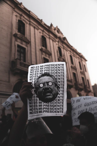 A protest scene with a hand holding a sign that repeats the phrase 'NO ES MI PRESIDENTE' alongside a stylized image of a man's face. The background features an old, grand building with arched windows and a crowd of people holding various signs, suggesting a demonstration in an urban environment.