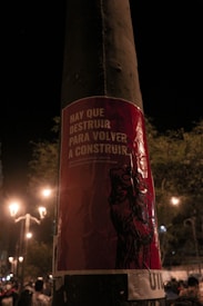 A poster with red and white text is affixed to a streetlight pole at night. The poster reads 'HAY QUE DESTRUIR PARA VOLVER A CONSTRUIR.' It features an illustration of a raised fist. The background shows streetlights illuminating a dark street with trees and some people visible in the distance.