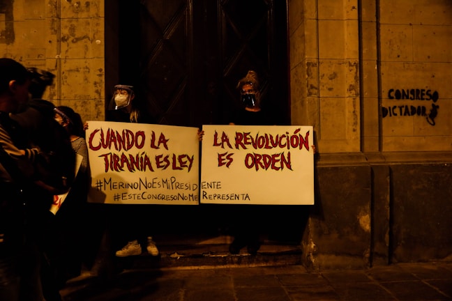 Several people are gathered in a dimly lit area holding protest signs in Spanish. The signs translate to messages that challenge tyranny and call for revolution as order. One person is wearing a face shield. There is graffiti on the wall that reads 'CONGRESO DICTADOR'. The mood suggests a political protest.