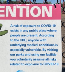 A sign attached to a chain-link fence provides a warning about the risk of exposure to COVID-19 in public places. It includes a message from the CDC highlighting the vulnerability of individuals with underlying medical conditions. The sign uses blue text on a white background with a prominent pink section at the top featuring the word 'ATTENTION'. In the background, a playground structure is partially visible.