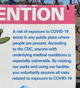 A sign attached to a chain-link fence provides a warning about the risk of exposure to COVID-19 in public places. It includes a message from the CDC highlighting the vulnerability of individuals with underlying medical conditions. The sign uses blue text on a white background with a prominent pink section at the top featuring the word 'ATTENTION'. In the background, a playground structure is partially visible.