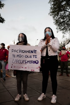 Two people stand side by side holding a sign. They wear face shields and casual clothing. The sign contains a message in Spanish addressing someone named Merino. Other people are present in the background, some wearing masks. Trees and a cloudy sky can be seen above them.