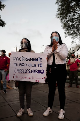 Two people stand side by side holding a sign. They wear face shields and casual clothing. The sign contains a message in Spanish addressing someone named Merino. Other people are present in the background, some wearing masks. Trees and a cloudy sky can be seen above them.