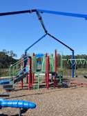 A brightly colored playground set featuring slides, climbing structures, and monkey bars. Red, blue, and green components contrast with the natural surroundings. The playground is situated on a wood chip-covered surface under a clear blue sky with trees in the background.
