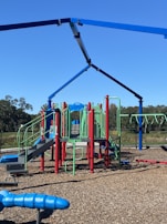 A brightly colored playground set featuring slides, climbing structures, and monkey bars. Red, blue, and green components contrast with the natural surroundings. The playground is situated on a wood chip-covered surface under a clear blue sky with trees in the background.