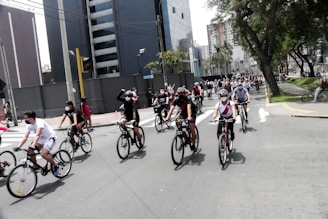 A large group of people riding bicycles along an urban street, with buildings and trees lining the road. Many of the cyclists are wearing helmets and face masks, and they appear to be part of an organized event or rally. The atmosphere seems lively and the weather is likely warm, given their attire.