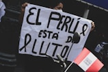 A person is holding a sign with large bold letters that read 'EL PERU ESTÁ DE LUTO' next to a white ribbon, indicating mourning. A red and white flag, likely representing Peru, is attached to a bicycle handlebar in the foreground.