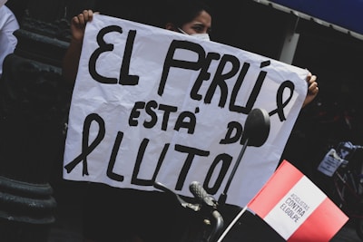 A person is holding a sign with large bold letters that read 'EL PERU ESTÁ DE LUTO' next to a white ribbon, indicating mourning. A red and white flag, likely representing Peru, is attached to a bicycle handlebar in the foreground.
