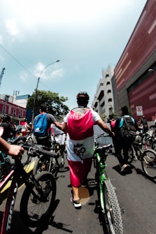 A group of people is participating in a street event, walking with bicycles on a city road. The scene includes a diverse crowd, with some individuals wearing backpacks and casual clothing. A person in the center is draped in a flag, suggesting a national or cultural event. Surrounding them are urban buildings, a clear sky, and a street sign.