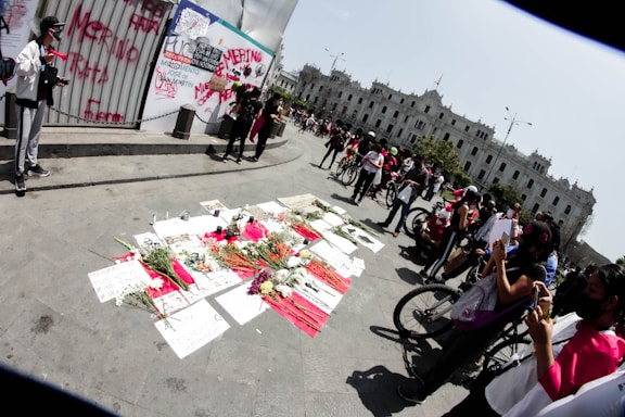 A street scene depicts a gathering of people near a makeshift memorial with flowers and written messages placed on the ground. Bicycles are scattered around, and several individuals are using their phones to take pictures. A large white building is visible in the background, while graffiti is present on a nearby wall.