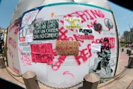 A wall covered in various protest posters and graffiti with messages in Spanish. The signs include phrases like 'Merino Golpista' and 'Hoy mi pais esta de luto por un crimen que no cometio.' The graffiti is made with different colors such as red, black, and green. There's also an image of a statue head and people in the background of a city square.