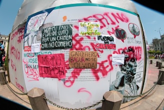 A wall covered in various protest posters and graffiti with messages in Spanish. The signs include phrases like 'Merino Golpista' and 'Hoy mi pais esta de luto por un crimen que no cometio.' The graffiti is made with different colors such as red, black, and green. There's also an image of a statue head and people in the background of a city square.