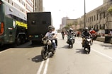 Several motorcyclists ride down a city street lined with buildings. A large police vehicle is parked on the left side. The scene is bustling with people walking around, some dressed in red shirts. The street is broad, bordered by urban architecture with some trees visible in the background.