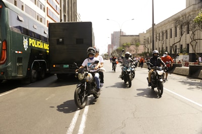 Several motorcyclists ride down a city street lined with buildings. A large police vehicle is parked on the left side. The scene is bustling with people walking around, some dressed in red shirts. The street is broad, bordered by urban architecture with some trees visible in the background.