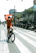 A cyclist wearing an orange shirt and a face mask holds a red and white flag while riding down a city street. The street is lined with onlookers and is bordered by tall buildings with some greenery. Overhead, there are streetlights and a clear sky.