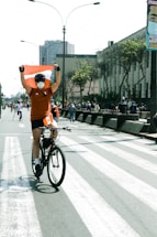 A colorful bicyclist with glowing lights rides through a city street, carrying vibrant two-sided banner and trailing double-sided flag banners.