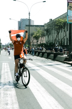 A cyclist wearing an orange shirt and a face mask holds a red and white flag while riding down a city street. The street is lined with onlookers and is bordered by tall buildings with some greenery. Overhead, there are streetlights and a clear sky.