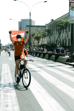 A colorful bicyclist with glowing lights rides through a city street, carrying vibrant two-sided banner and trailing double-sided flag banners.