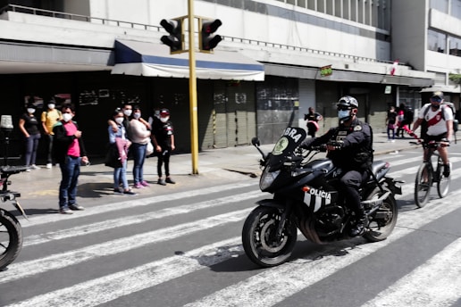 A police officer wearing a face mask rides a black motorcycle with visible 'Policia' markings across a crosswalk. Simultaneously, cyclists and pedestrians, all wearing face masks, are in the vicinity. The backdrop features a city street with shuttered storefronts and a yellow traffic light post.