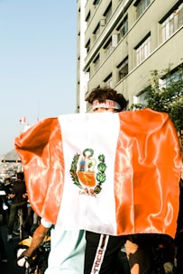 A person is wearing a large flag over their shoulders that features a coat of arms, possibly from Peru. The setting appears to be a busy urban street with a tall building and several people visible in the background. Light reflects off the flag, indicating it's a bright day.