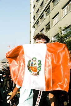 A person is wearing a large flag over their shoulders that features a coat of arms, possibly from Peru. The setting appears to be a busy urban street with a tall building and several people visible in the background. Light reflects off the flag, indicating it's a bright day.